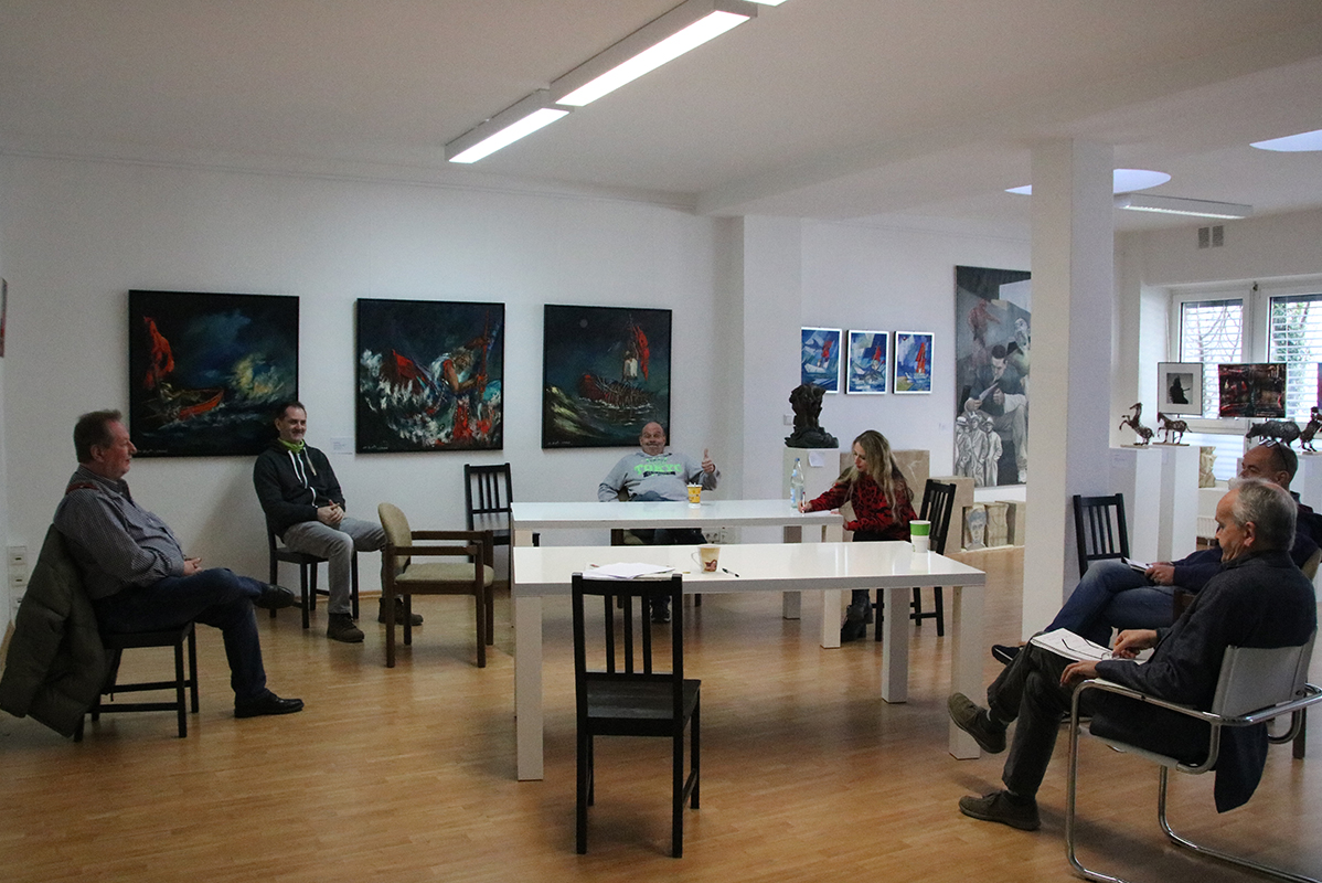 6 Menschen sitzen in einer Kunstgalerie im 1,5-Meter-Abstand bei einer Besprechung