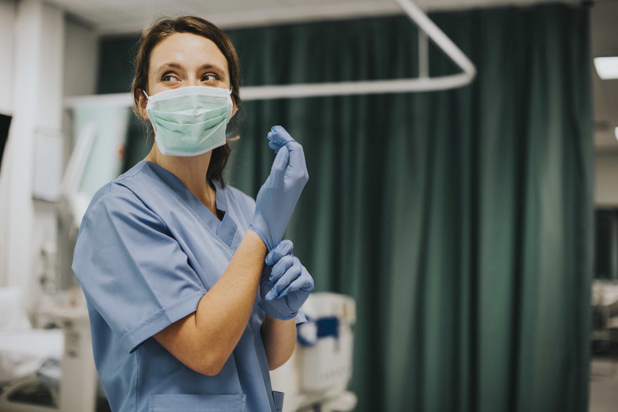 Krankenschwester mit Mund-Nase-Schutz zieht sich lächelnd Einmalhandschuh über die Hand